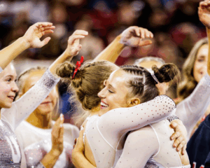 Utah gymnasts celebrate on the sideline, with Ella Zirbes smiling as she hugs a teammate in a white, rhinestone-studded leotard. Other teammates surround them with arms raised and hands mid-cheer, capturing a moment of excitement and camaraderie during a meet.