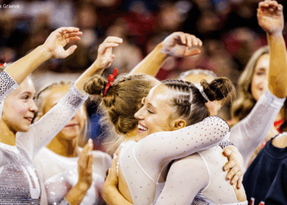Utah gymnasts celebrate on the sideline, with Ella Zirbes smiling as she hugs a teammate in a white, rhinestone-studded leotard. Other teammates surround them with arms raised and hands mid-cheer, capturing a moment of excitement and camaraderie during a meet.