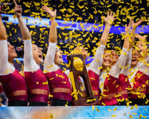The 2026 Oklahoma gymnastics team hold their hands high as gold confeti falls and they hold the NCAA championship trophy in the center of the photo.