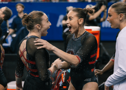 Two Stanford gymnasts, Ana Barbosu and Levi Jung-Ruivivar, react with excitement on the competition floor, facing each other with wide smiles and open mouths as they celebrate a routine. Both wear black leotards with red accents and have their hair in tight buns, while a teammate stands nearby watching the moment unfold.