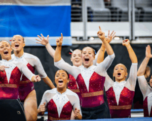 Members of the Oklahoma gymnastics team celebrate during the 2026 NCAA Championships, wearing red and white leotards. Several gymnasts stand together with arms raised and mouths open in excitement, cheering toward the competition floor as a routine concludes. The arena seating and competition setup are visible in the background.