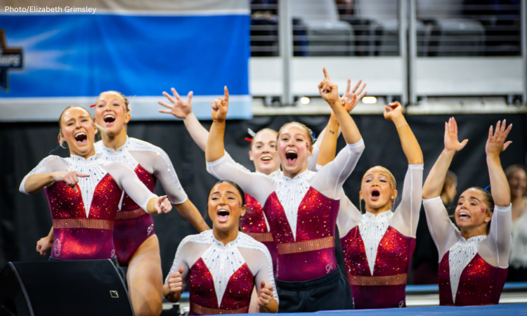 Members of the Oklahoma gymnastics team celebrate during the 2026 NCAA Championships, wearing red and white leotards. Several gymnasts stand together with arms raised and mouths open in excitement, cheering toward the competition floor as a routine concludes. The arena seating and competition setup are visible in the background.