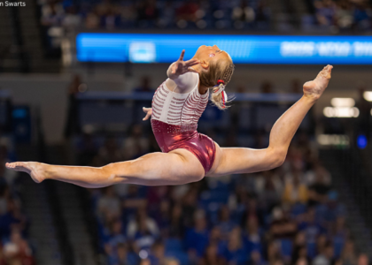 Ella Murphy does a ring leap on beam with her legs in a split, her back leg bent, and her head back.