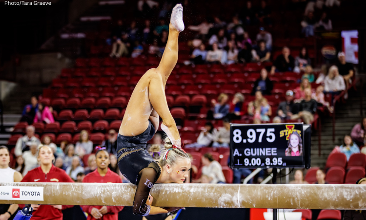 Finley Weldon poses on beam in a chin stand with both arms wrapped under the beam, one leg extended vertically upwards, and the other bent towards her head. 