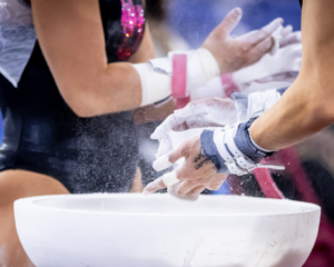 Two gymnasts stand by a chalk bucket chalking their grips.