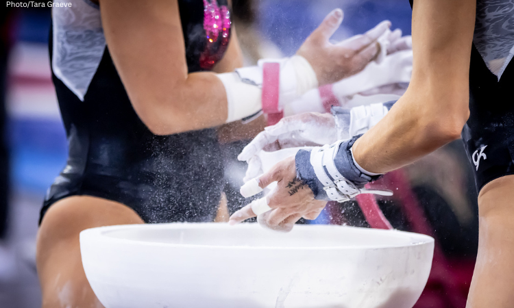 Two gymnasts stand by a chalk bucket chalking their grips.