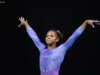 Gymnast Christina Shelton stands on the competition floor in a lavender long-sleeve leotard with intricate geometric patterns and rhinestone details. With both arms raised in a poised dance pose, she looks ahead with a focused expression against a dark arena backdrop.