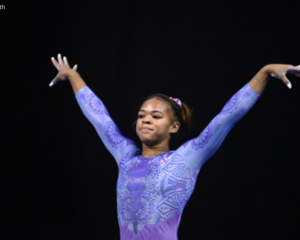 Gymnast Christina Shelton stands on the competition floor in a lavender long-sleeve leotard with intricate geometric patterns and rhinestone details. With both arms raised in a poised dance pose, she looks ahead with a focused expression against a dark arena backdrop.