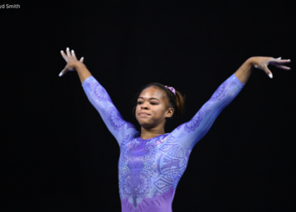 Gymnast Christina Shelton stands on the competition floor in a lavender long-sleeve leotard with intricate geometric patterns and rhinestone details. With both arms raised in a poised dance pose, she looks ahead with a focused expression against a dark arena backdrop.