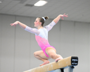 Caroline Moreau of Texas Dreams balances in a low crouch on the balance beam during competition, wearing a pink-and-white leotard with her arms extended gracefully to either side. The background is softly blurred, drawing focus to her poised beam choreography.