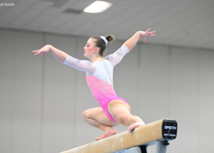 Caroline Moreau of Texas Dreams balances in a low crouch on the balance beam during competition, wearing a pink-and-white leotard with her arms extended gracefully to either side. The background is softly blurred, drawing focus to her poised beam choreography.