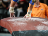 A close-up view of a gymnastics vault table covered in white chalk marks. A gymnast’s taped wrists and chalked hands make contact with the table during a vault, while the background is blurred, showing spectators and an official in an orange shirt.