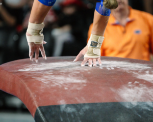 A close-up view of a gymnastics vault table covered in white chalk marks. A gymnast’s taped wrists and chalked hands make contact with the table during a vault, while the background is blurred, showing spectators and an official in an orange shirt.