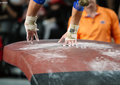 A close-up view of a gymnastics vault table covered in white chalk marks. A gymnast’s taped wrists and chalked hands make contact with the table during a vault, while the background is blurred, showing spectators and an official in an orange shirt.