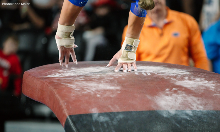 A close-up view of a gymnastics vault table covered in white chalk marks. A gymnast’s taped wrists and chalked hands make contact with the table during a vault, while the background is blurred, showing spectators and an official in an orange shirt.