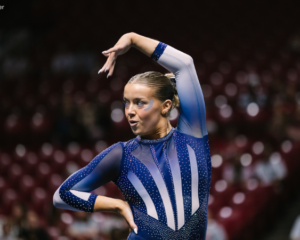 New Hampshire gymnast Anne Challman poses during her floor exercise routine, with one arm arched overhead and the other resting at her waist. She wears a blue rhinestone leotard with sheer sleeves and white accents, and blue eye makeup. Blurred arena seating and spectators fill the background.
