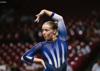 New Hampshire gymnast Anne Challman poses during her floor exercise routine, with one arm arched overhead and the other resting at her waist. She wears a blue rhinestone leotard with sheer sleeves and white accents, and blue eye makeup. Blurred arena seating and spectators fill the background.