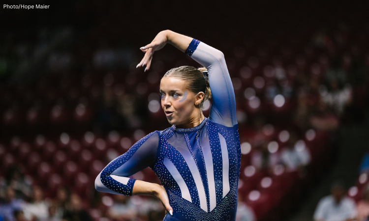 New Hampshire gymnast Anne Challman poses during her floor exercise routine, with one arm arched overhead and the other resting at her waist. She wears a blue rhinestone leotard with sheer sleeves and white accents, and blue eye makeup. Blurred arena seating and spectators fill the background.