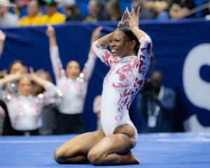Jamison Sears of Alabama kneels on the floor at the end of her routine, smiling as she holds her hands above her head in a celebratory pose. She wears a white leotard with crimson swirl detailing, while her cheering teammates and a blurred arena crowd fill the background.