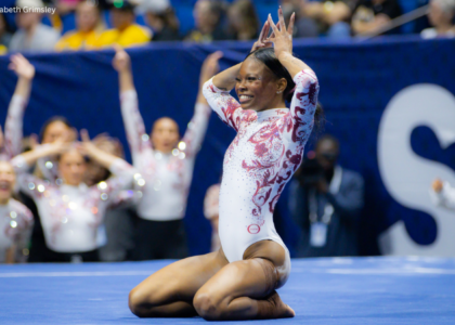 Jamison Sears of Alabama kneels on the floor at the end of her routine, smiling as she holds her hands above her head in a celebratory pose. She wears a white leotard with crimson swirl detailing, while her cheering teammates and a blurred arena crowd fill the background.