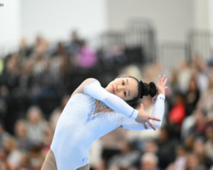 Jayla Hang performs a dance element during a floor exercise routine, leaning sideways with her arms extended in an expressive pose. She wears a light blue, long-sleeve leotard with rhinestone detailing, while a blurred crowd of spectators fills the background. Photo by Lloyd Smith.