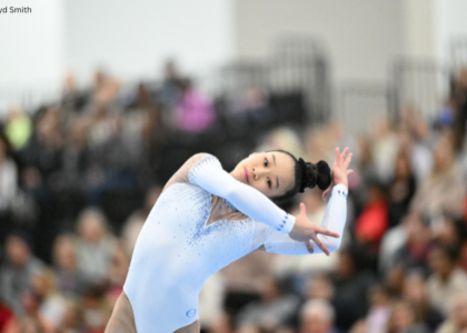 Jayla Hang performs a dance element during a floor exercise routine, leaning sideways with her arms extended in an expressive pose. She wears a light blue, long-sleeve leotard with rhinestone detailing, while a blurred crowd of spectators fills the background. Photo by Lloyd Smith.