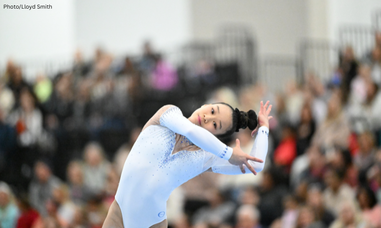 Jayla Hang performs a dance element during a floor exercise routine, leaning sideways with her arms extended in an expressive pose. She wears a light blue, long-sleeve leotard with rhinestone detailing, while a blurred crowd of spectators fills the background. Photo by Lloyd Smith.
