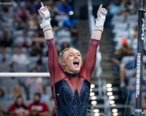 Minnesota gymnast Gianna Gerdes celebrates after her bar routine with both arms raised overhead and her mouth open in a cheer. She wears a maroon and black long-sleeve leotard with sparkling details and white grips, while the uneven bars and a blurred arena crowd fill the background.