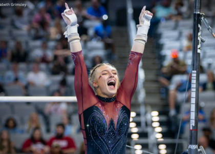 Minnesota gymnast Gianna Gerdes celebrates after her bar routine with both arms raised overhead and her mouth open in a cheer. She wears a maroon and black long-sleeve leotard with sparkling details and white grips, while the uneven bars and a blurred arena crowd fill the background.