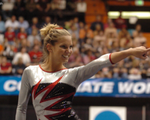 Georgia gymnast Abby Stack smiles and points toward the crowd while standing on the competition floor during the 2006 NCAA National Championships. She wears a silver, black, and red long-sleeve leotard, with the arena crowd blurred in the background.