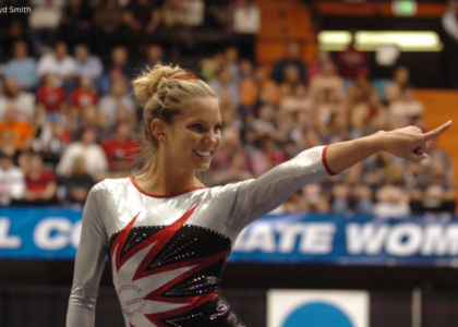 Georgia gymnast Abby Stack smiles and points toward the crowd while standing on the competition floor during the 2006 NCAA National Championships. She wears a silver, black, and red long-sleeve leotard, with the arena crowd blurred in the background.