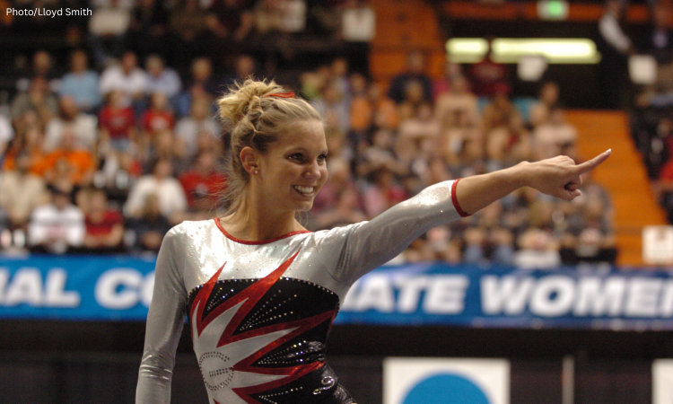 Georgia gymnast Abby Stack smiles and points toward the crowd while standing on the competition floor during the 2006 NCAA National Championships. She wears a silver, black, and red long-sleeve leotard, with the arena crowd blurred in the background.